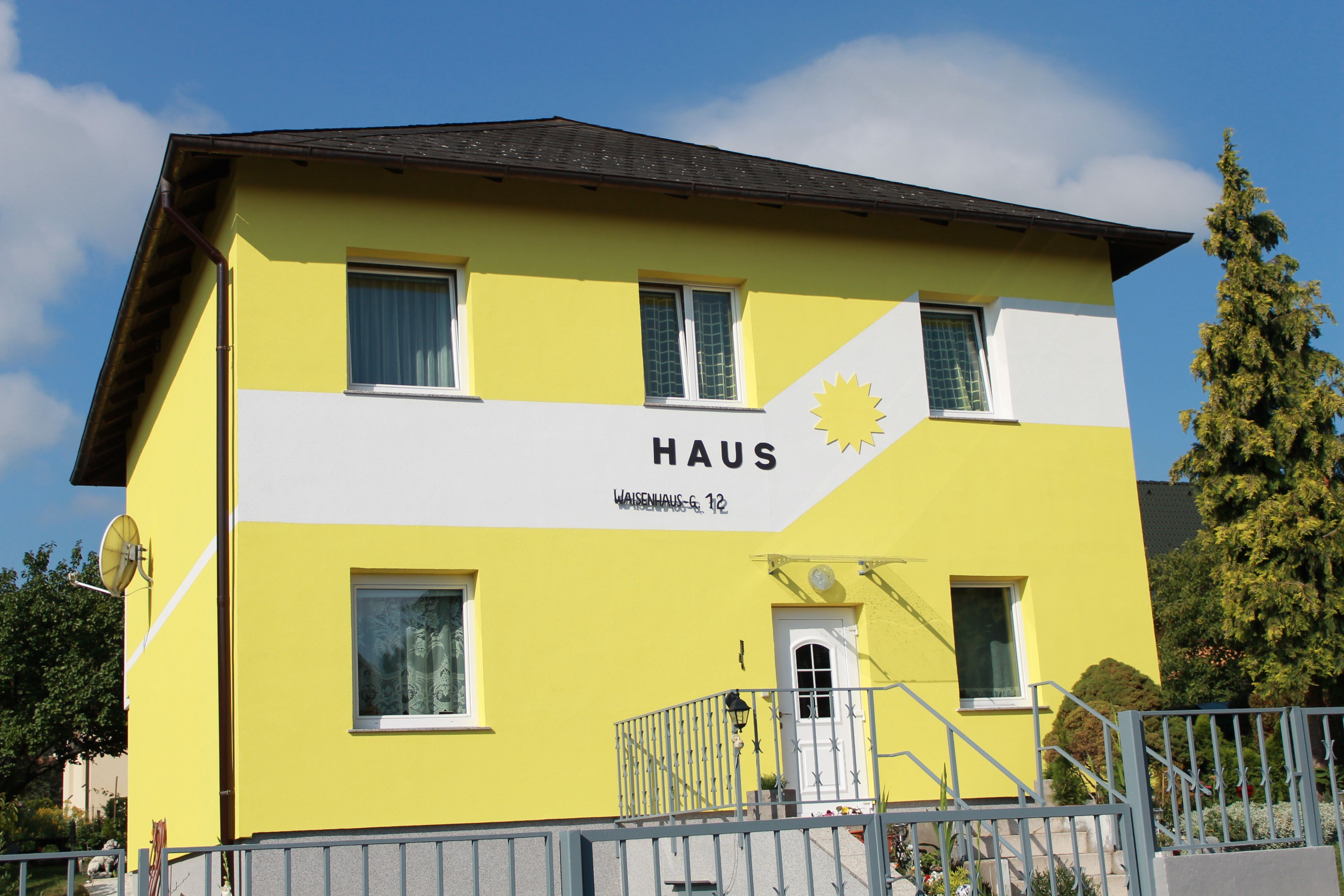 Gelbes Haus mit der Aufschrift 'Haus' und einer Sonne auf der Fassade.