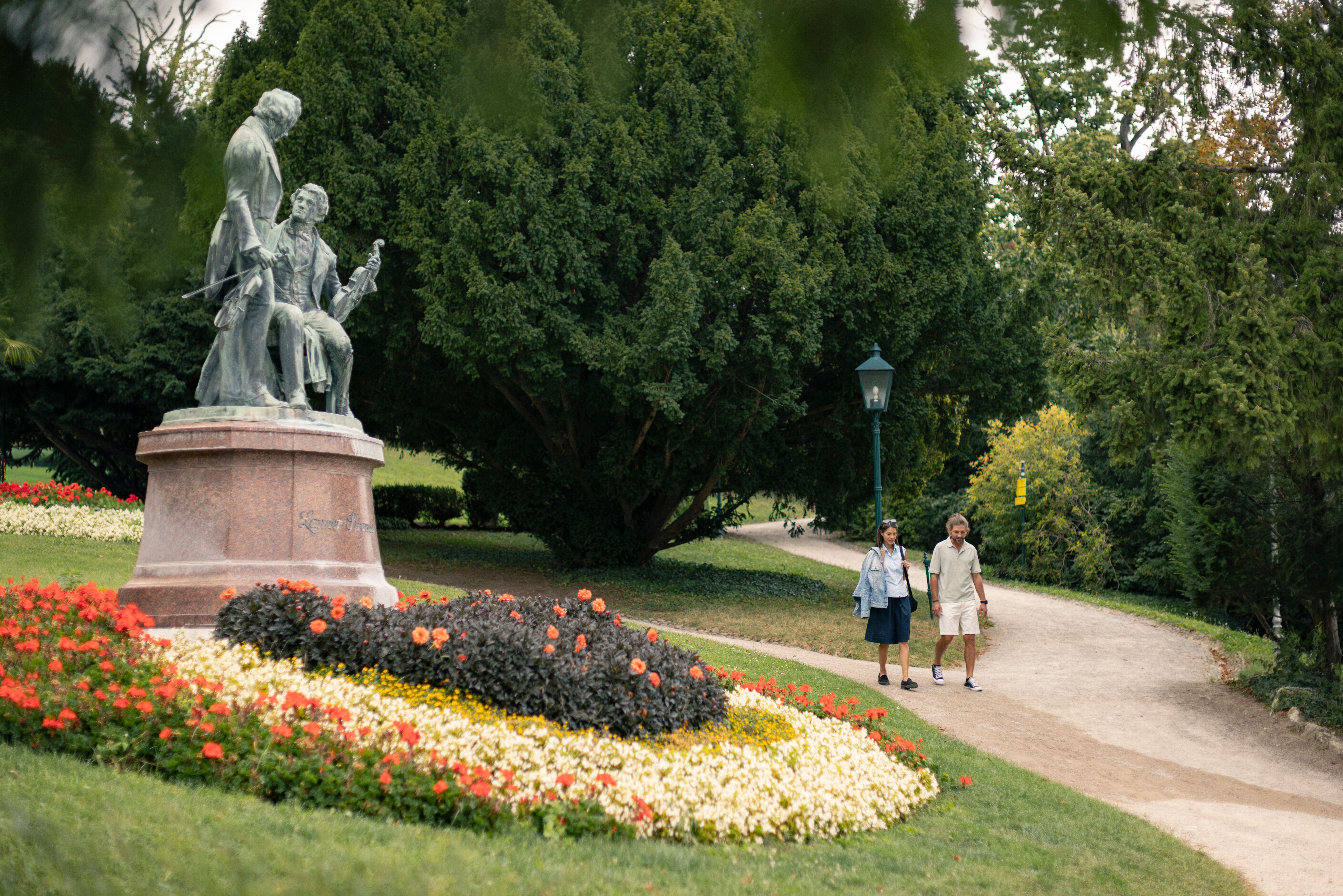 Eine Frau und ein Mann schlendern im Kurpark Baden am Lanner Strauß Denkmal vorbei. Vor ihnen erstreckt sich ein großes Blumenbeet mit roten, gelben und weißen Blumen. Im Hintergrund sind Bäume des Parks sichtbar.