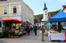 Ein belebter Bauernmarkt mit St&auml;nden und Besuchern vor einem gelben Geb&auml;ude und einer Kirche im Hintergrund.