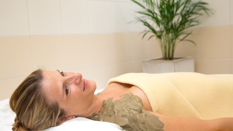 Frau entspannt mit Schwefelschlammpackung auf der Schulter, eingewickelt in eine Decke, im Wellnessbereich mit Pflanze im Hintergrund.