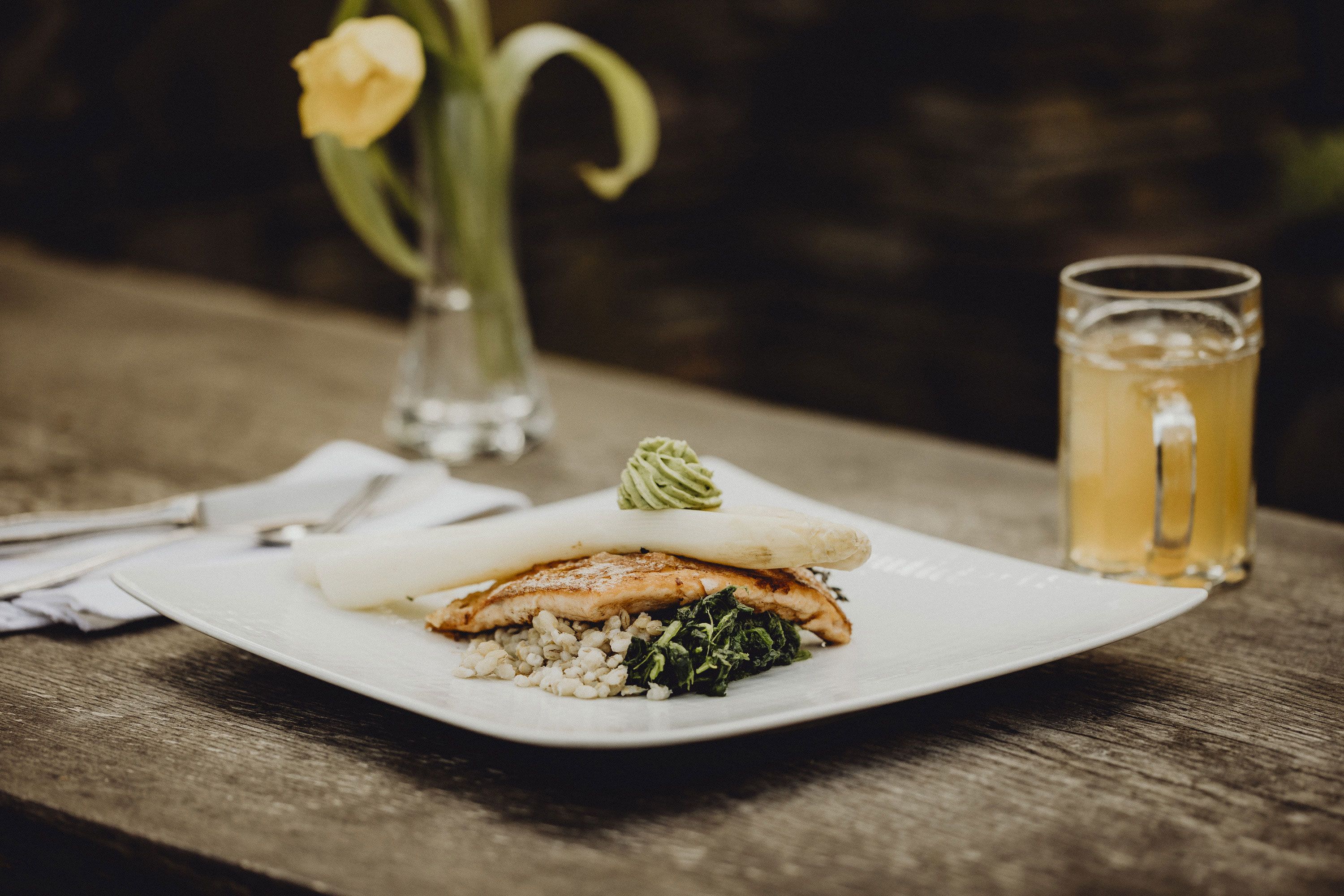 Gourmetgericht mit Spargel, Fisch und Gemüse auf Holztisch, gelbe Blume im Hintergrund.