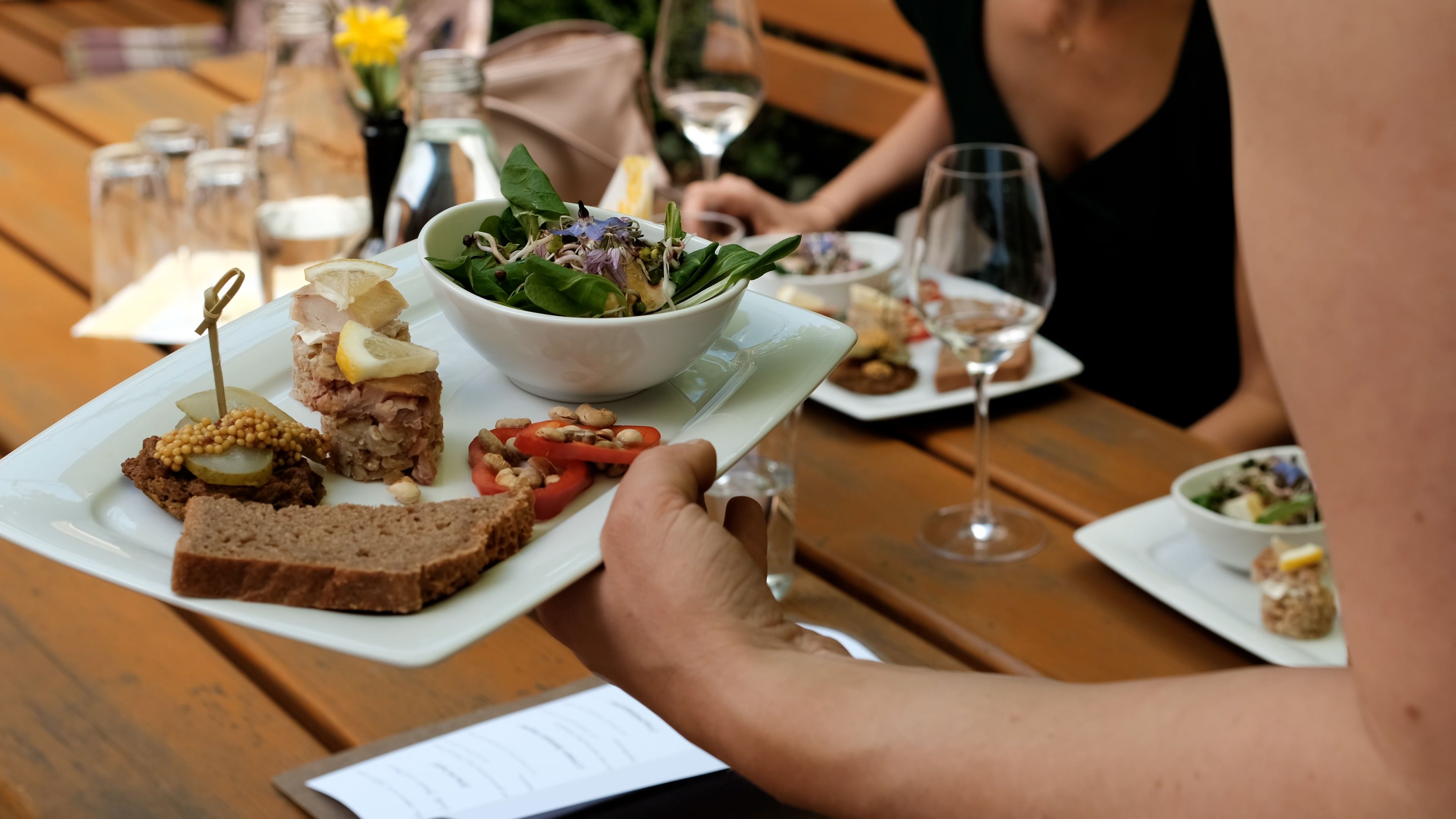 Auf einem Tisch im Freien steht eine Vorspeisenvariation mit frischem Salat und Brot als Beilage. Eine Hand hält einen Teller über dem sommerlich gedeckten Heurigentisch. Das helle Tageslicht und die lockere Atmosphäre vermitteln ein sommerliches Genusserlebnis.