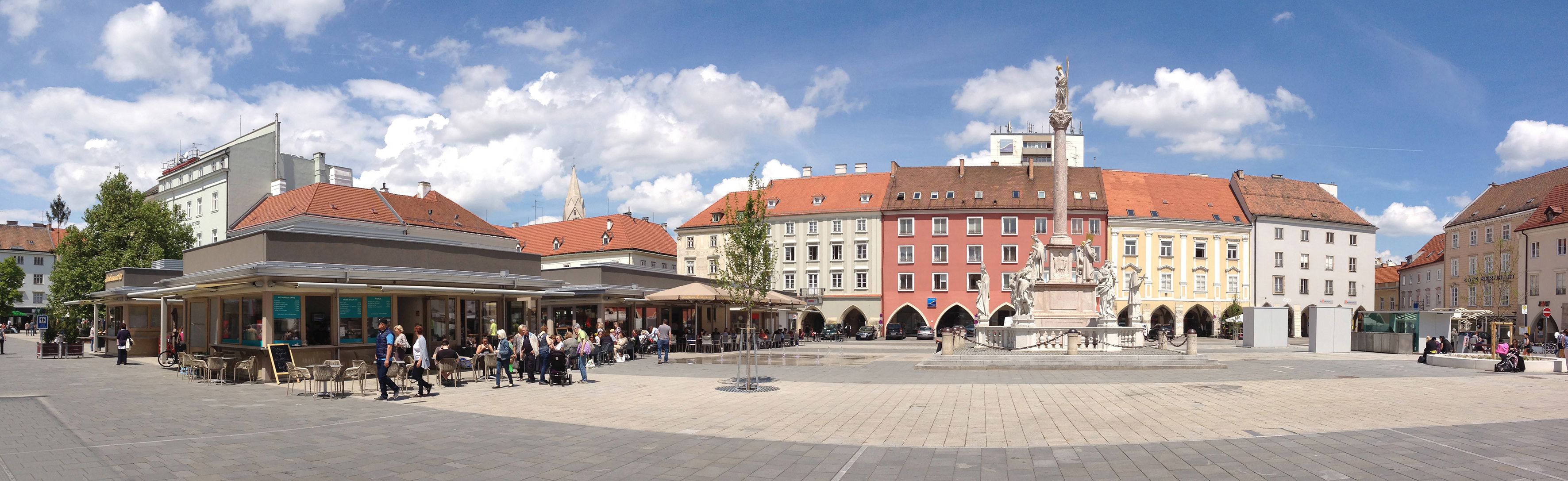 Panoramablick auf einen belebten Hauptplatz mit historischen Gebäuden und einer Statue in der Mitte.
