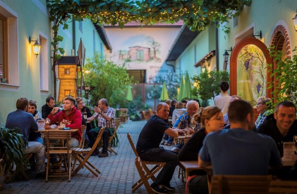 Menschen sitzen an Holztischen in einem gemütlichen Innenhof eines Weinguts, umgeben von Pflanzen und Lichterketten.