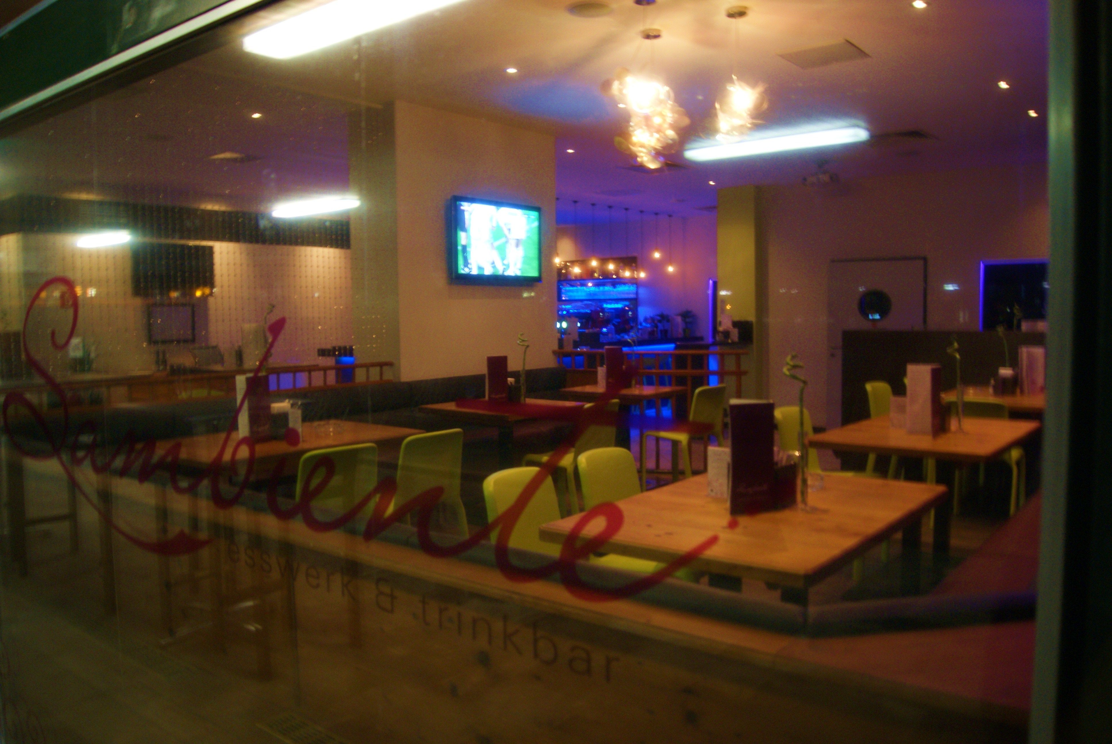 Innenansicht eines leeren Restaurants mit Holztischen und bunten Stühlen, Fernseher an der Wand und beleuchteter Bar im Hintergrund.