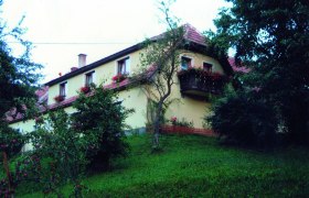 Ein gelbes Haus mit roten Dachziegeln und Blumenkästen an den Fenstern, umgeben von Bäumen und einem grünen Hang.