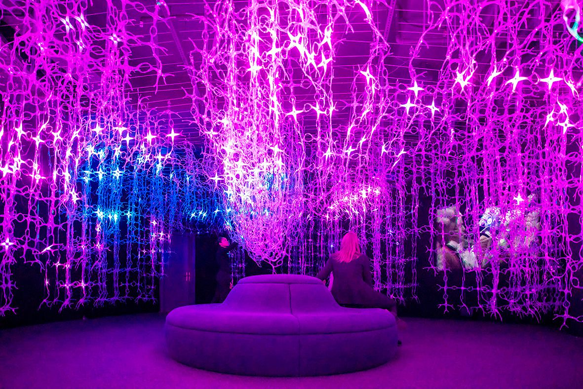 Ein Raum mit violetten und blauen Lichterketten, die von der Decke hängen. Eine Person sitzt auf einem runden Sofa.