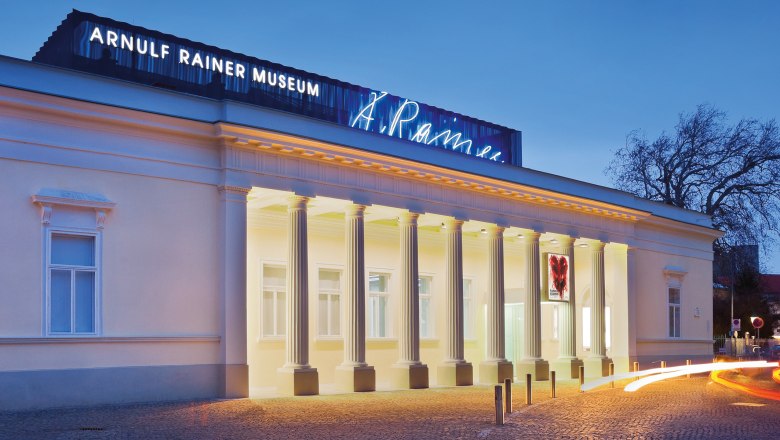 Außenansicht des Arnulf Rainer Museums bei Dämmerung mit beleuchteten Säulen und Leuchtschrift.