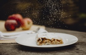 Apple strudel with powdered sugar on a plate, apples in the background.