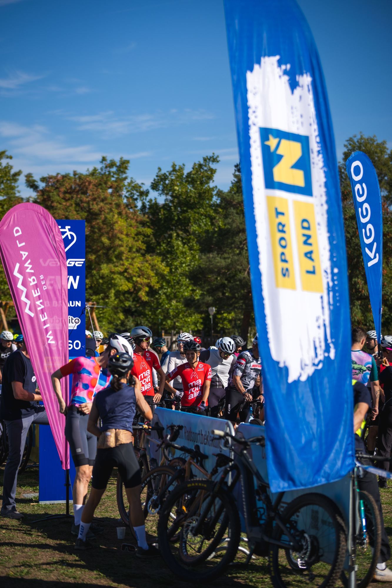 Gruppe von Radfahrerinnen und Radfahrern mit Gravelbikes zwischen bunten Veranstaltungsbannern unter freiem Himmel.