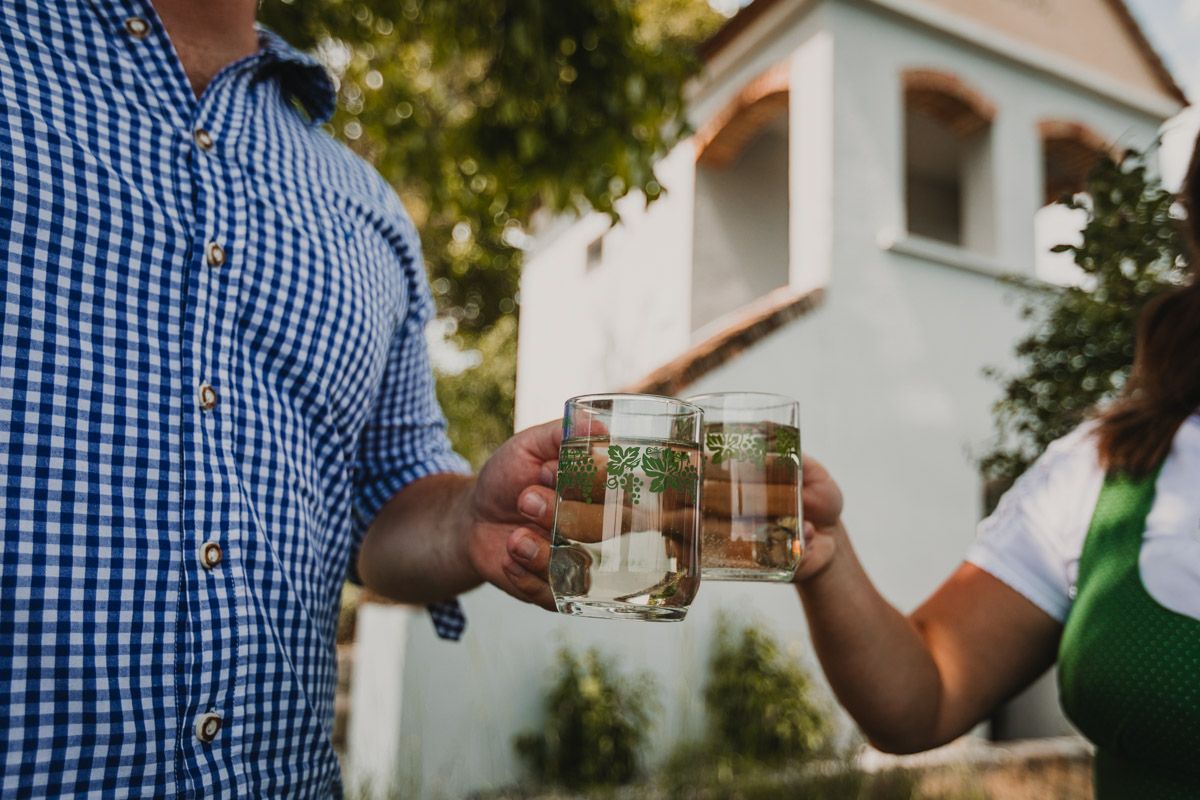 Eine Frau und ein Mann stoßen im Sommer mit Weißwein-Spritzer-Gläsern vor einem Gebäude im Weingarten an, der Mann trägt ein blau-weiß kariertes Hemd.