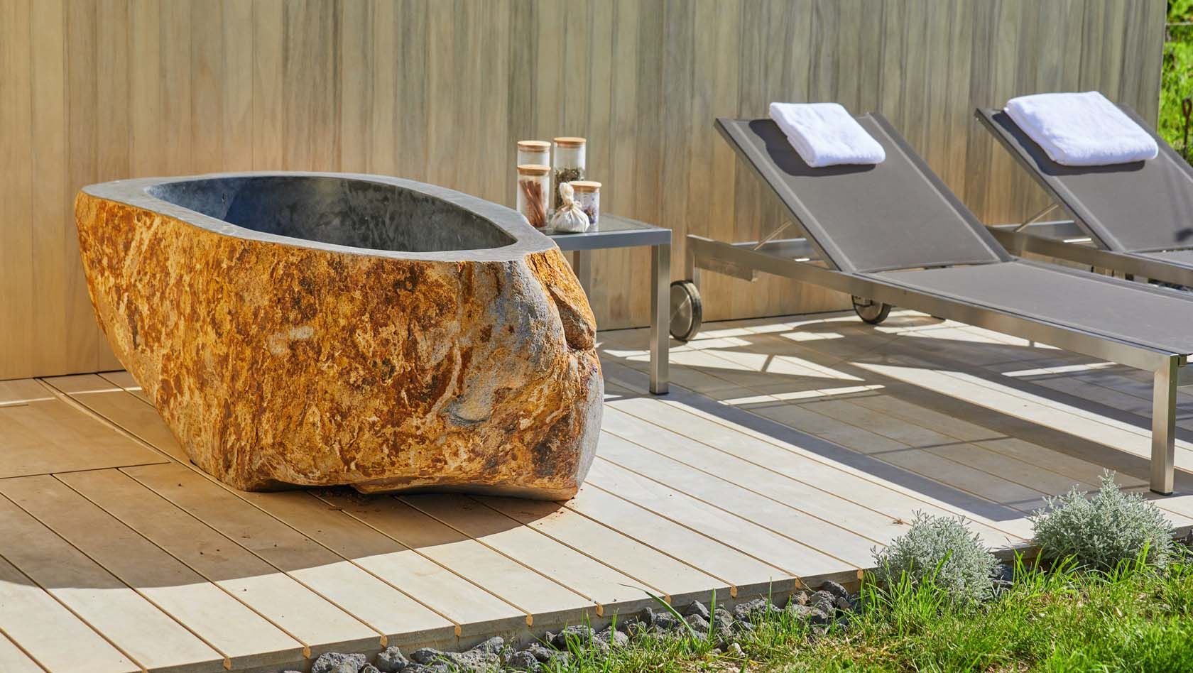Steinbadewanne und Liegestühle auf Holzterrasse im Freien.