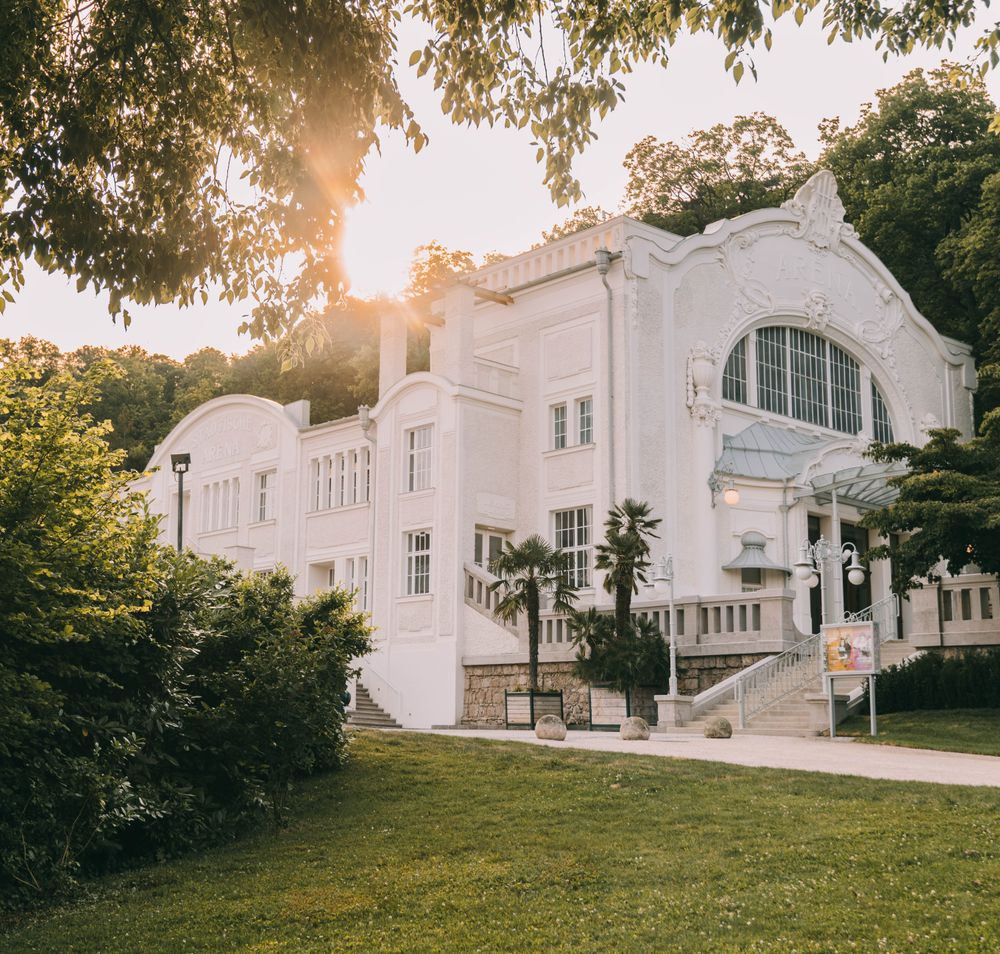 Inmitten der malerischen Kulisse des Wienerwaldes strahlt das historische Gebäude in sanften Pastellfarben. Die warmen Sonnenstrahlen tauchen die Umgebung in ein goldenes Licht und laden dazu ein, die kulturellen Veranstaltungen des Theaterfests zu genießen. Üppige Pflanzen und gepflegte Grünflächen umrahmen diesen charmanten Ort und schaffen eine einladende Atmosphäre.