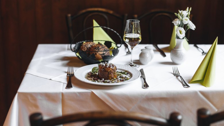 Ein gedeckter Tisch mit Tafelspitzsulz, Brot, einem Glas Weißwein und Blumen.