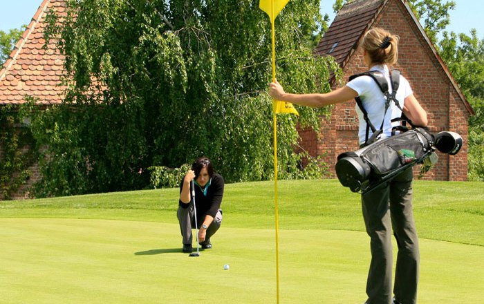 Zwei Frauen spielen Golf auf einem grünen Golfplatz, eine hält die Fahne, die andere zielt auf das Loch.