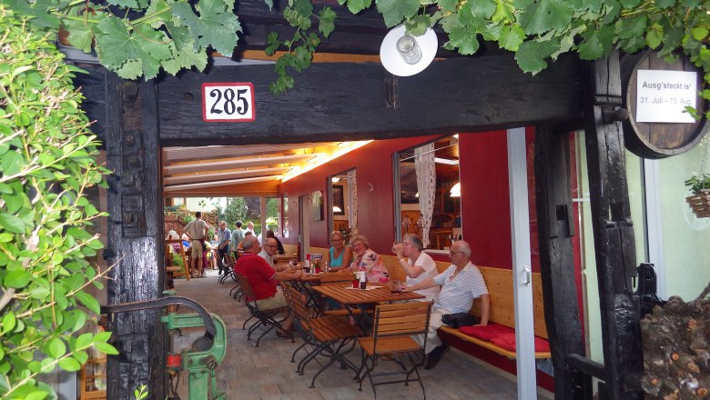 People sit at wooden tables in a covered outdoor area with vines.