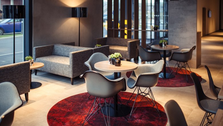 Moderne Hotellobby mit grauen Sofas, runden Holztischen und roten Teppichen.