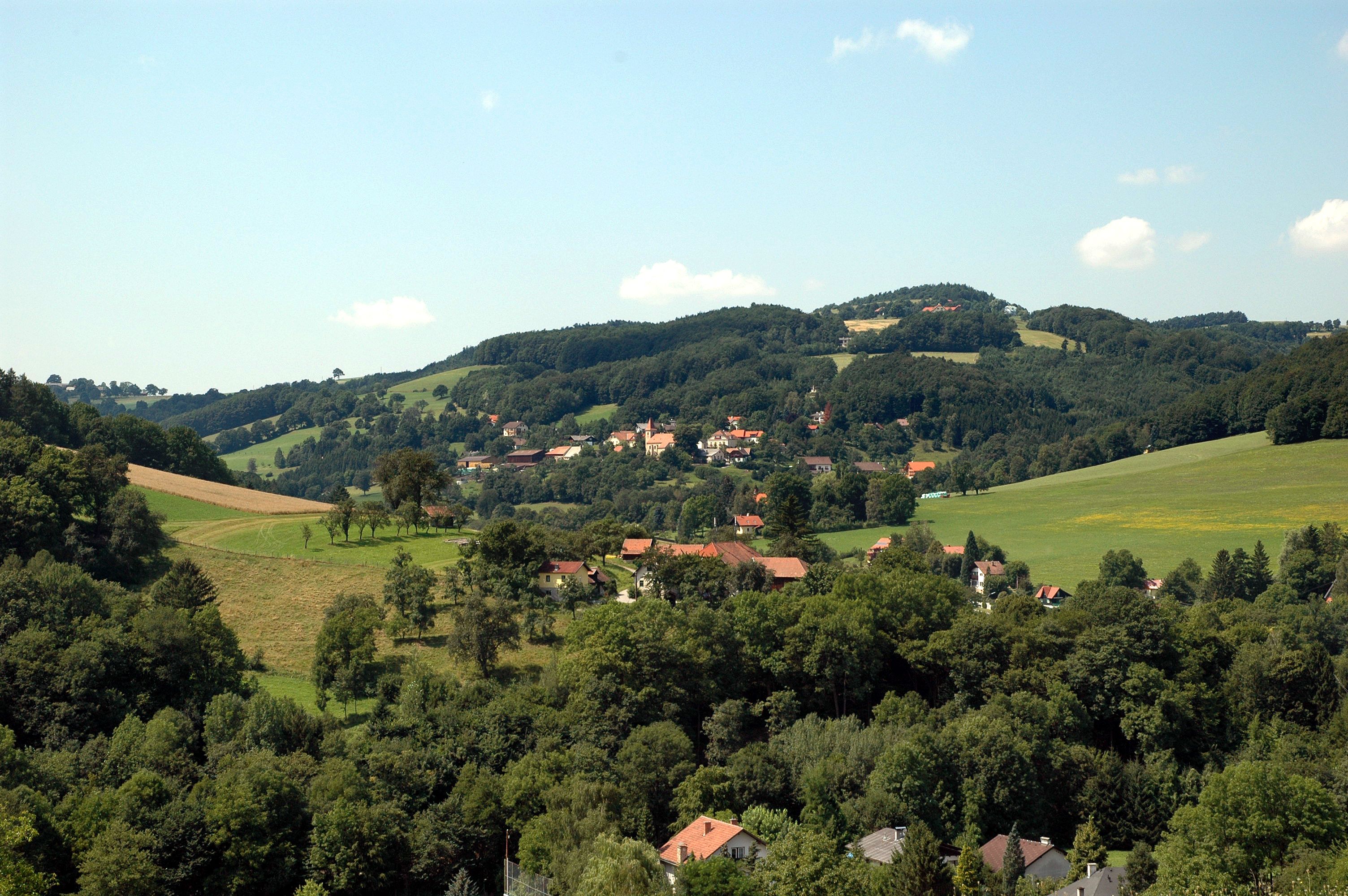 Landschaft mit Hügeln, Wäldern und einem Dorf im Hintergrund.