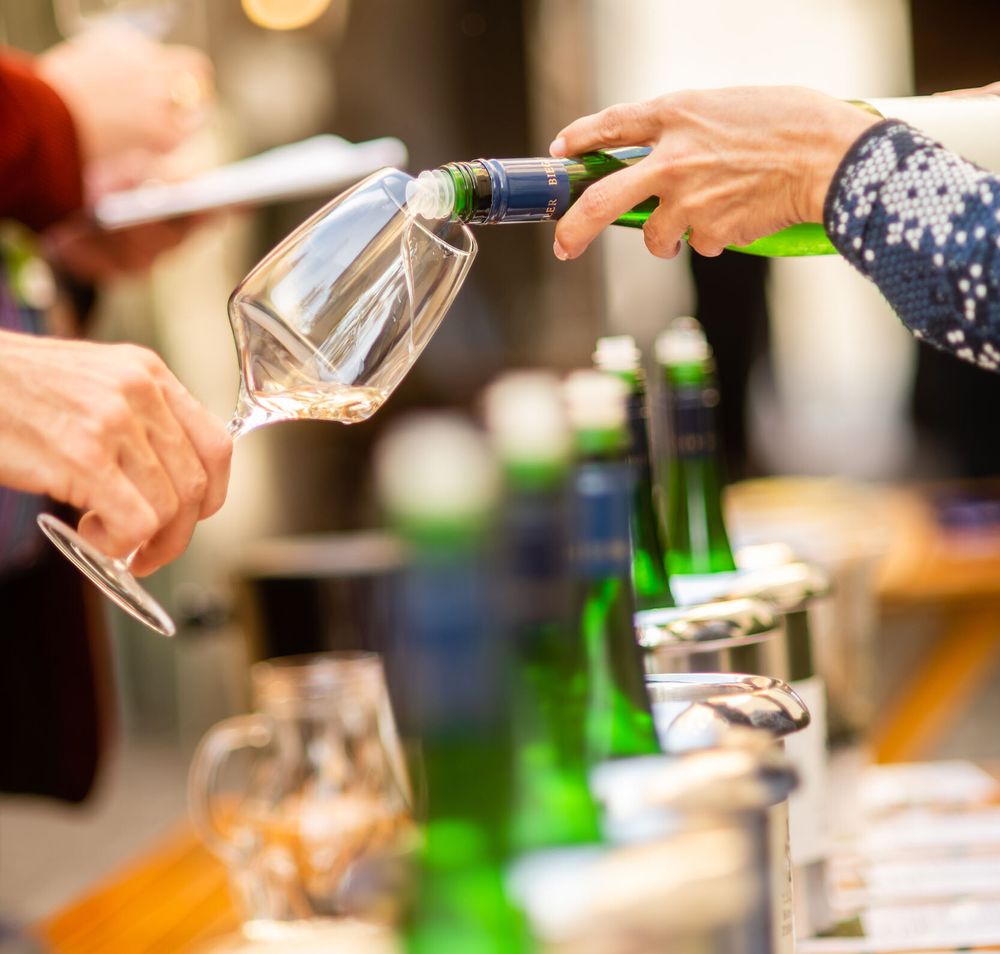Inmitten der malerischen Weinlandschaft wird ein Glas erlesener Wein eingeschenkt, während die Gäste in geselliger Runde anstoßen. Die sanften Hügel des Wienerwaldes umrahmen die Szenerie und laden dazu ein, die Aromen des Weins und die Schönheit der Natur zu genießen.