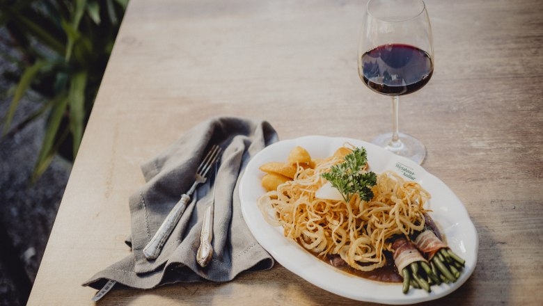 Ein Teller mit Zwiebelrostbraten, Kartoffeln und Speckbohnen, daneben ein Glas Rotwein auf einem Holztisch.