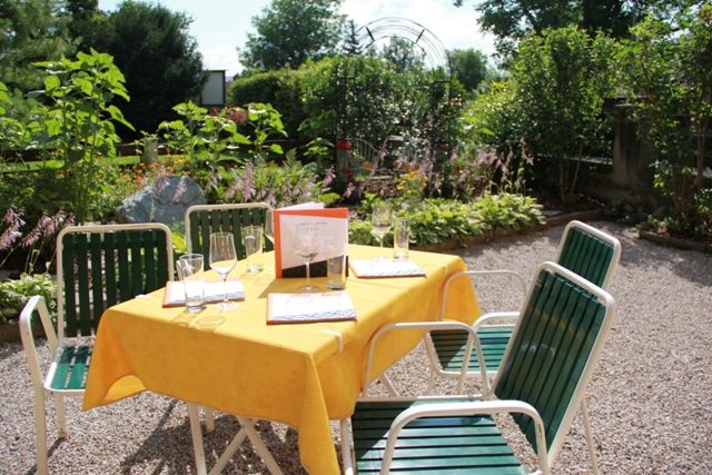 Ein sonniger Gastgarten mit einem Tisch, der mit gelber Tischdecke und Gläsern gedeckt ist, umgeben von grünen Pflanzen.
