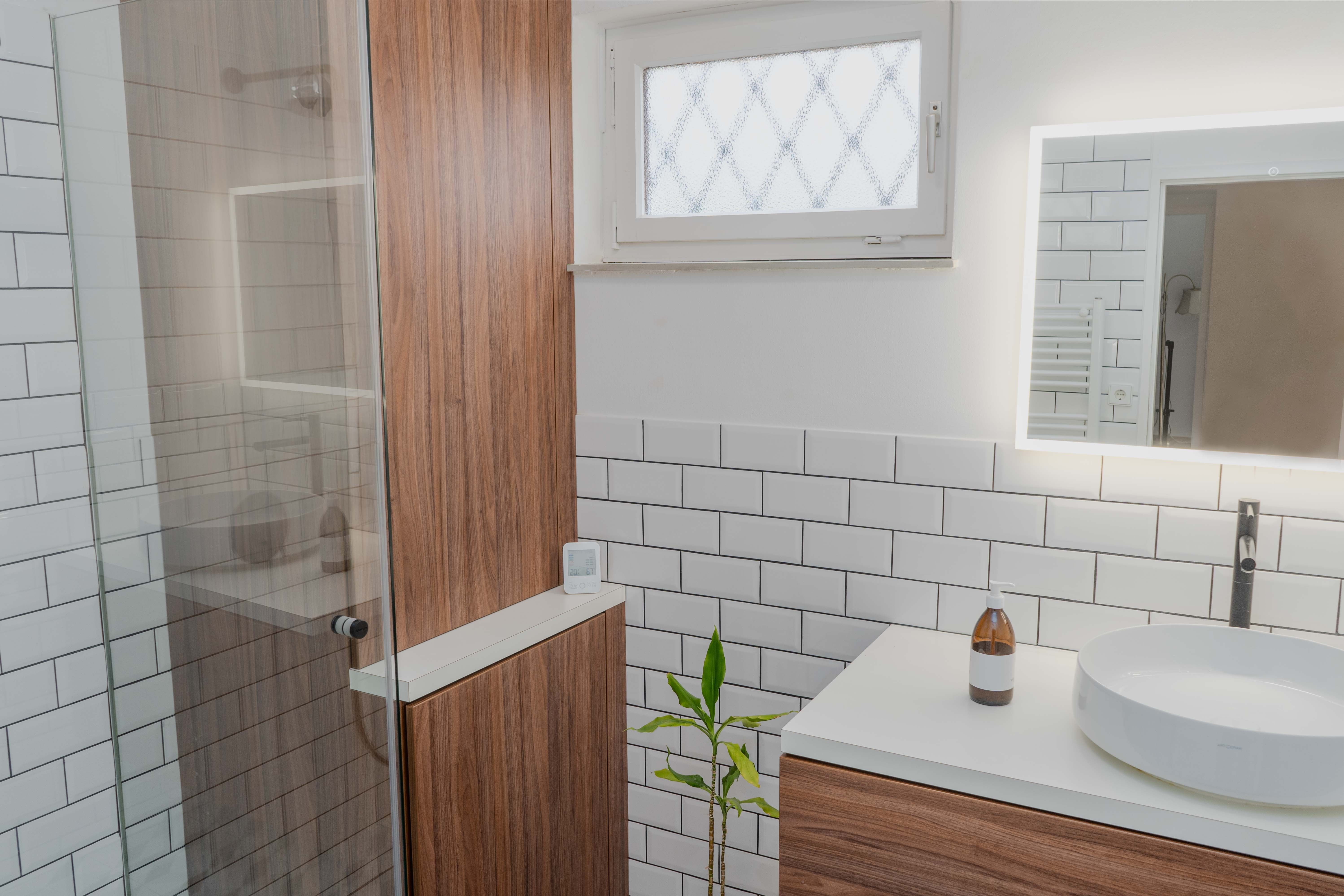 Modernes Badezimmer mit Holzschränken, weißer Fliesenwand, rundem Waschbecken und beleuchtetem Spiegel.