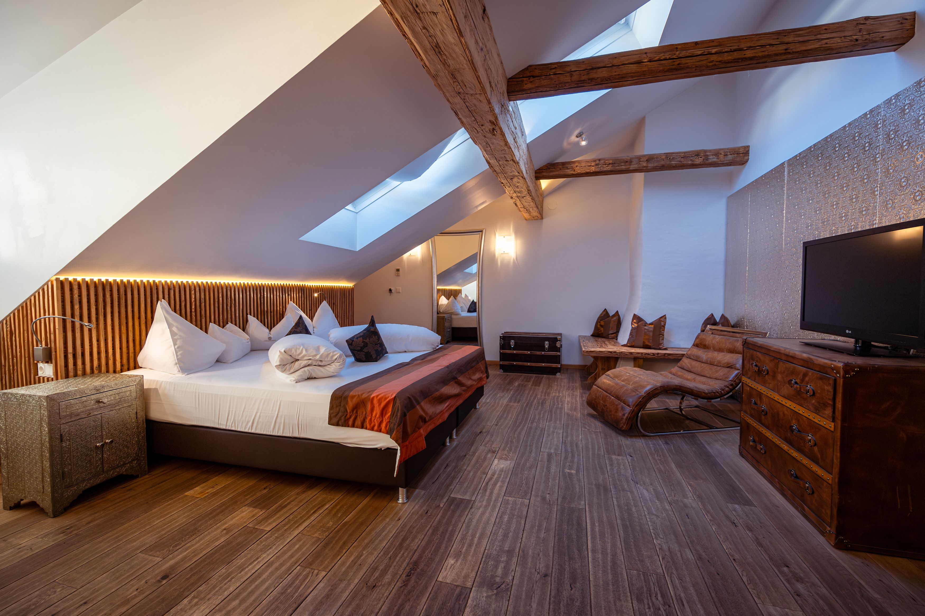 Gemütliches Hotelzimmer mit Holzbalken, Dachfenstern, großem Bett und Ledersessel.