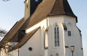 Totzenbach parish church with clock tower and pointed roof, surrounded by a fence, in daylight.