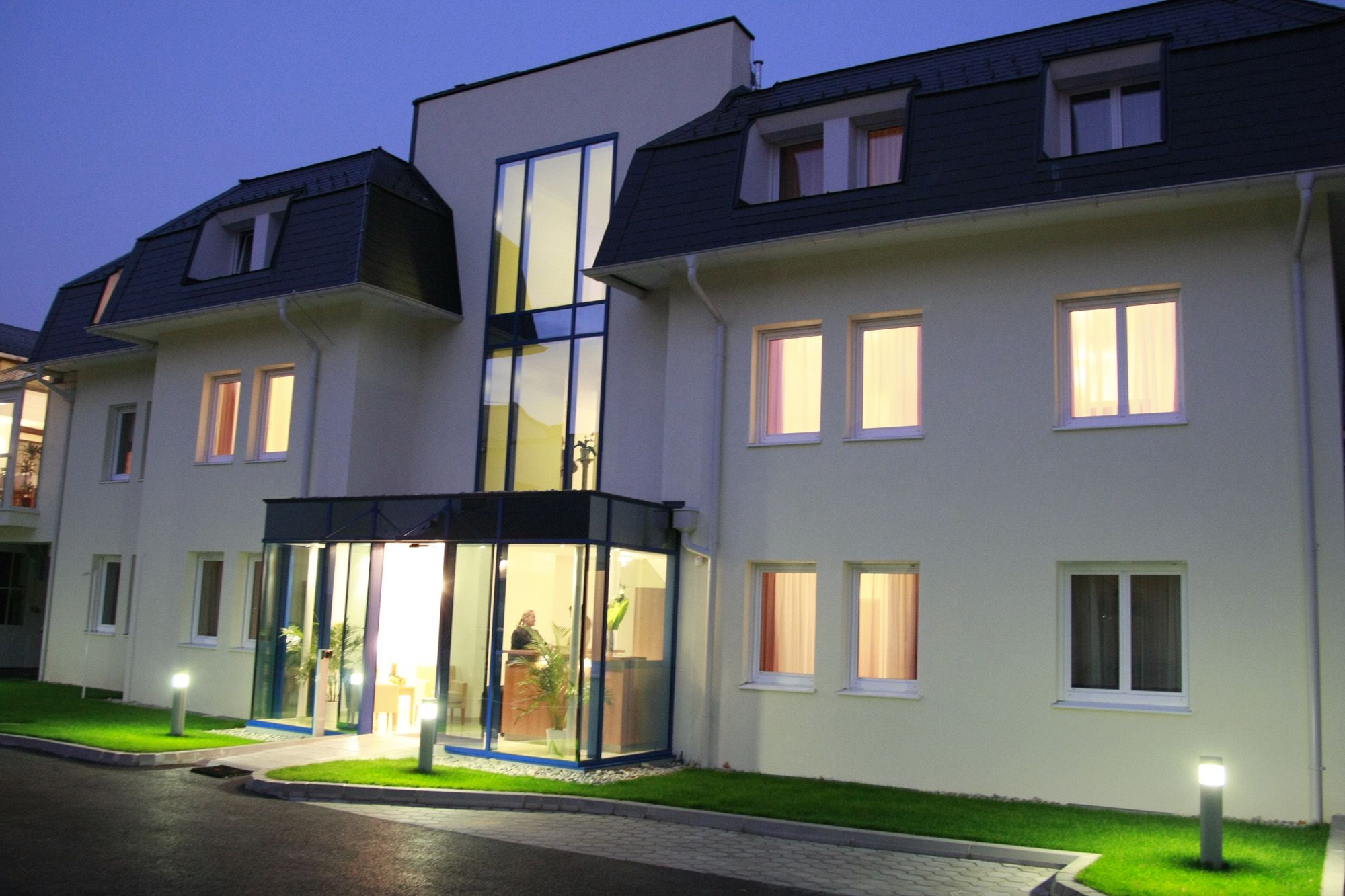 Eingangsbereich eines modernen Hotels bei Dämmerung mit beleuchteten Fenstern und Glasfassade.