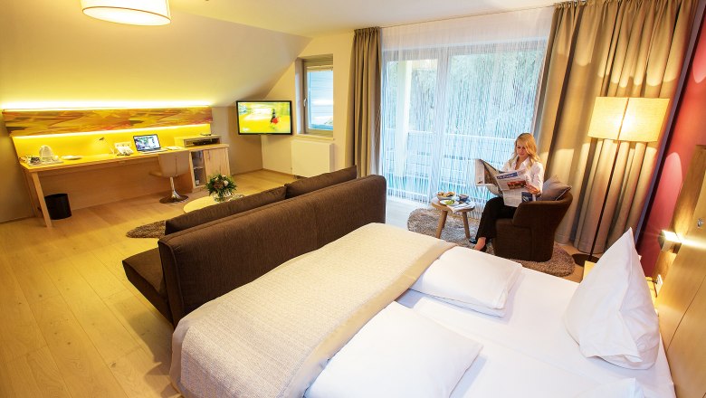 Modern hotel room with double bed, desk, TV and a woman reading the newspaper.