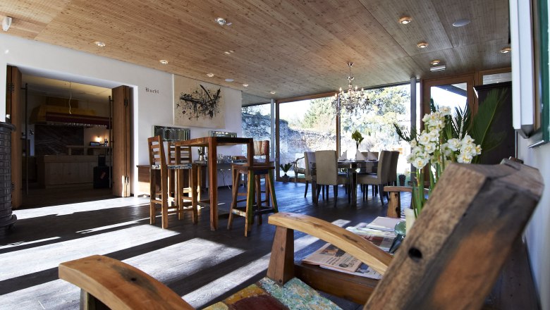 Ein moderner Frühstücksraum mit Holzmöbeln, großen Fenstern und einem Kronleuchter. Im Vordergrund ein Holzstuhl, im Hintergrund ein Esstisch.