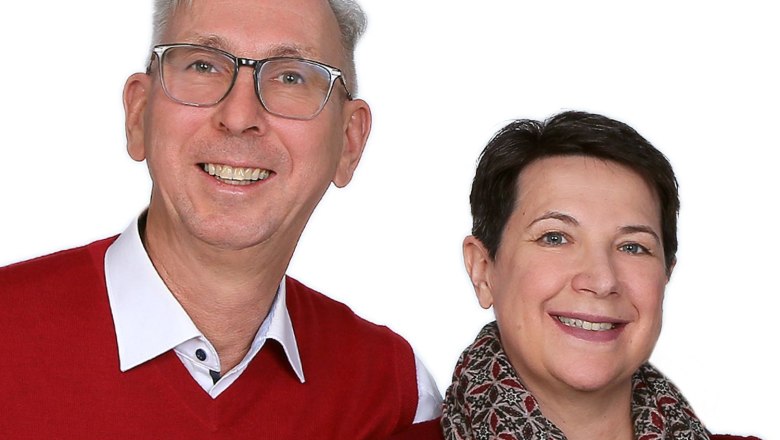 Ein Mann und eine Frau lächeln in die Kamera. Beide tragen rote Kleidung. Der Mann hat eine Brille, die Frau einen gemusterten Schal.