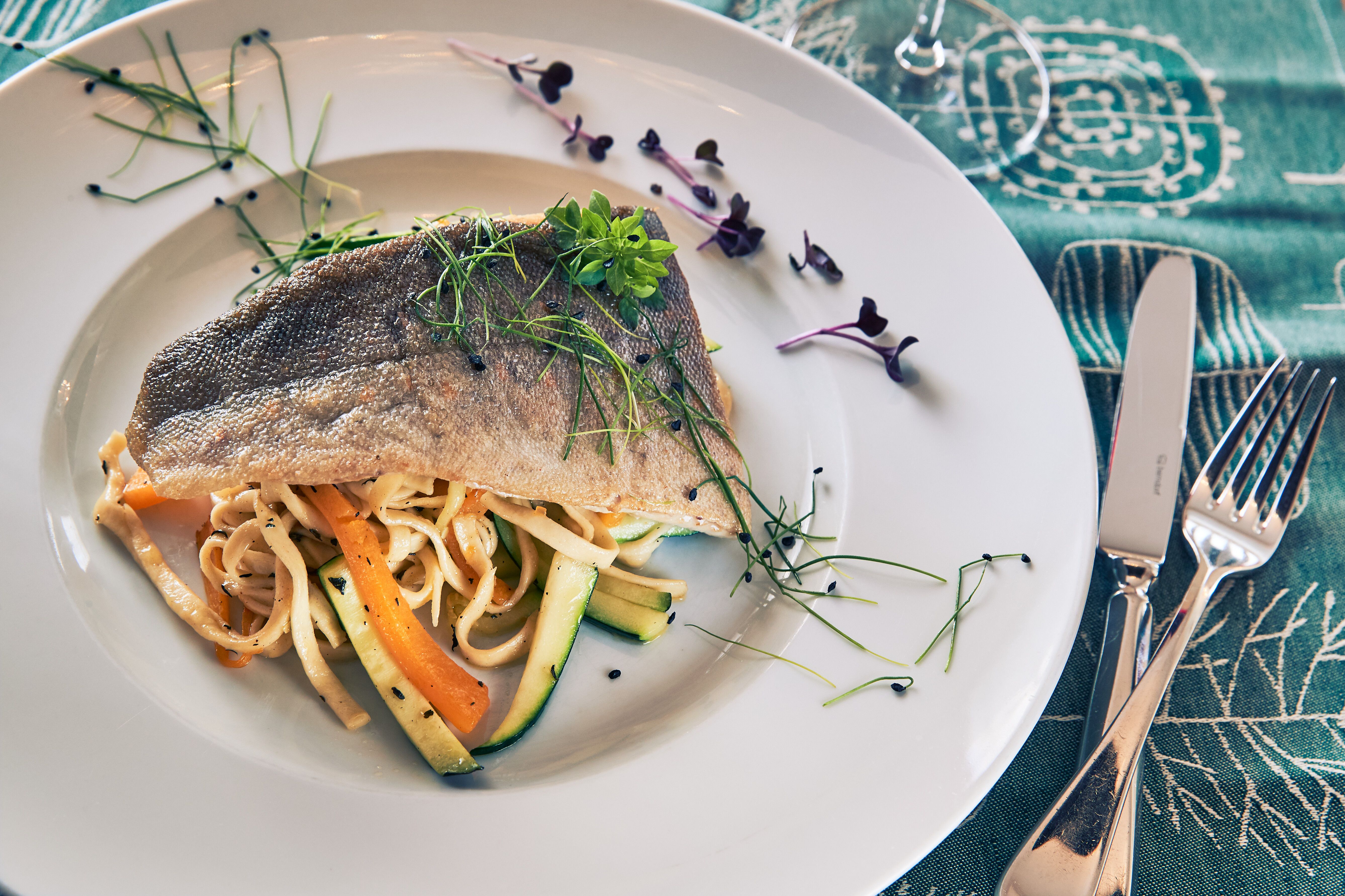 Ein köstliches Seesaiblingsfilet ruht auf einem Bett aus Kürbis-Zucchini-Nudeln, umgeben von frischen Kräutern. Diese harmonische Kombination verspricht ein unvergessliches Geschmackserlebnis inmitten der idyllischen Landschaft des Wienerwaldes.