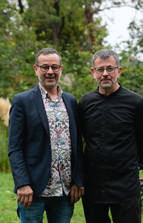 Die Brüder Franz und Karl Rosenbauch stehen nebeneinander in ihrem großen Garten, umgeben von Bäumen und Grün, beide lächelnd.