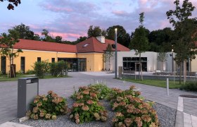 Modernes Gebäude mit gelber Fassade und rotem Dach, umgeben von Pflanzen und Bäumen, bei Sonnenuntergang.