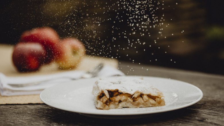 Apfelstrudel mit Puderzucker auf Teller, Äpfel im Hintergrund.