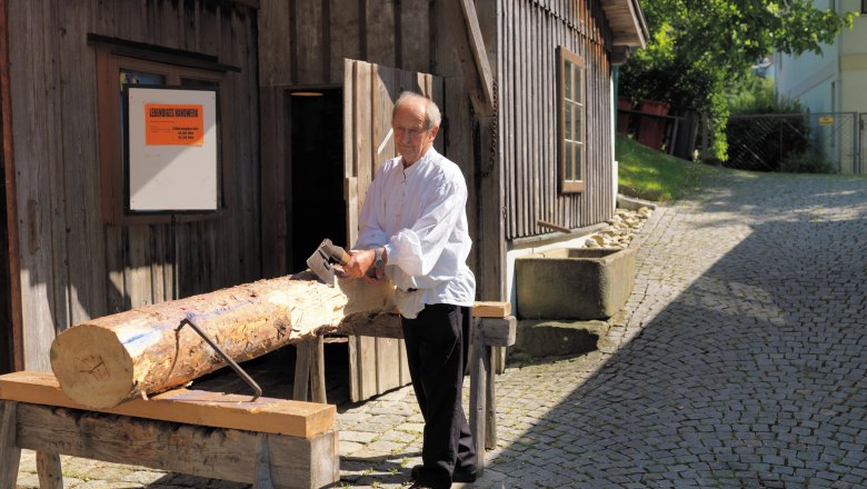 A man is working on a tree trunk with an axe in front of a wooden house.