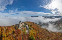 Luftaufnahme der Araburg im Herbst: Die Burg ist von orange- und gelb gefärbtem Wald umgeben, eine dichte Nebeldecke liegt über dem Tal, während oberhalb der Burg strahlender Sonnenschein den Himmel erhellt.