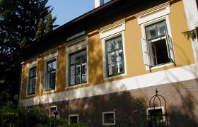 Yellow house with open windows and garden in the foreground.