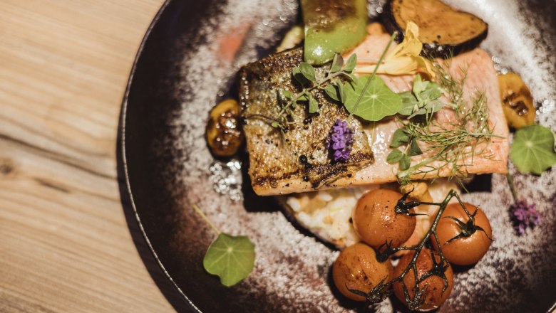 Lachsforellenfilet auf Grammelrisotto mit Gemüse und Kräutern auf einem Teller.