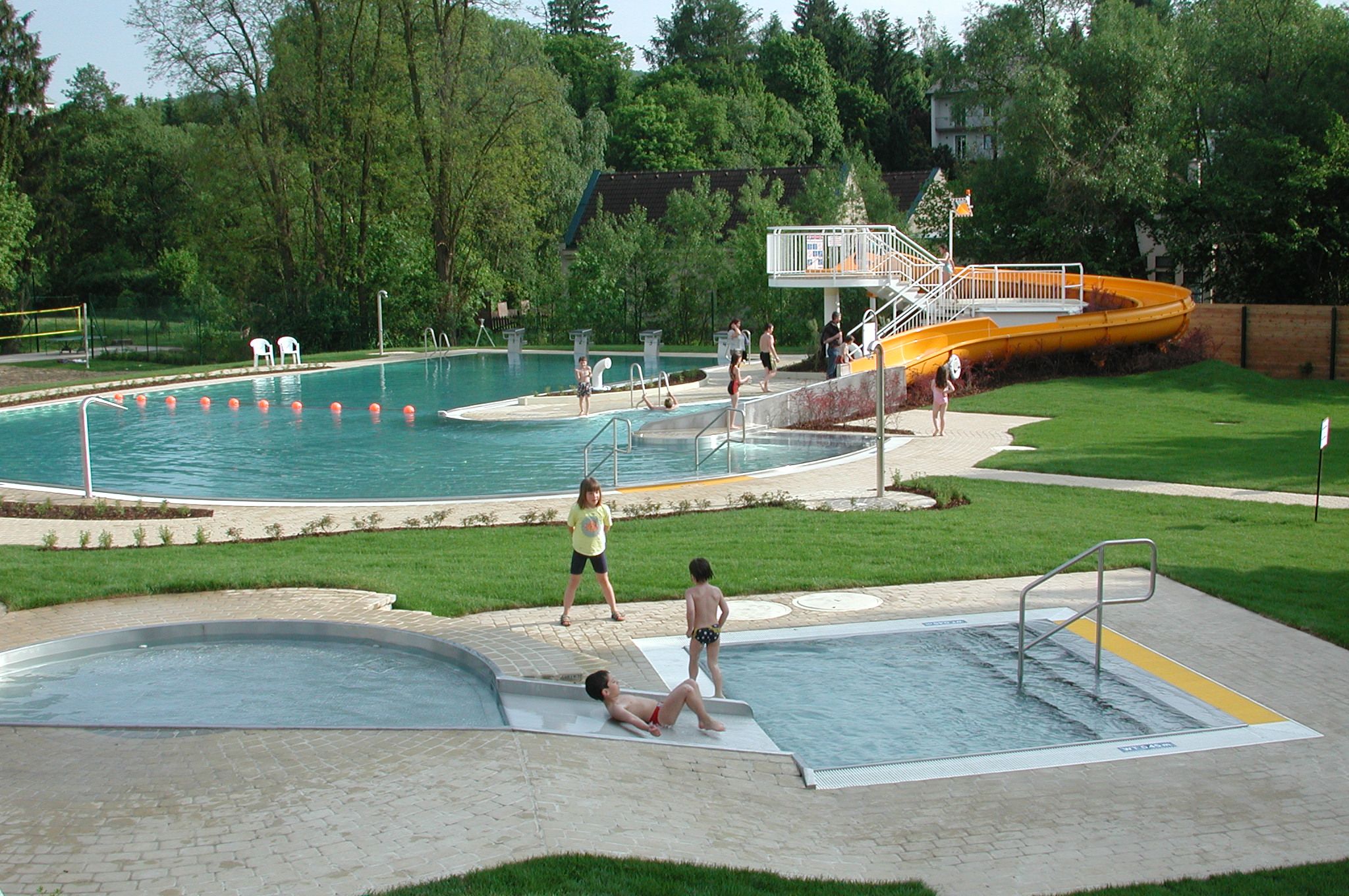 Freibad mit Kinderbecken und Rutsche, umgeben von Bäumen und Rasenflächen.