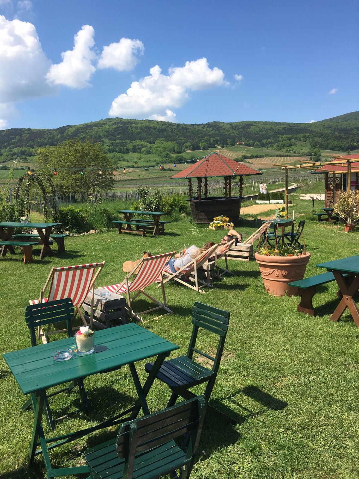 Garten mit Liegestühlen und Tischen, im Hintergrund Hügel und blauer Himmel.