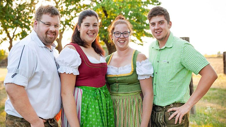 Vier Personen in Tracht stehen lächelnd im Freien auf einer grünen Wiese, im Hintergrund ist ein Baum.