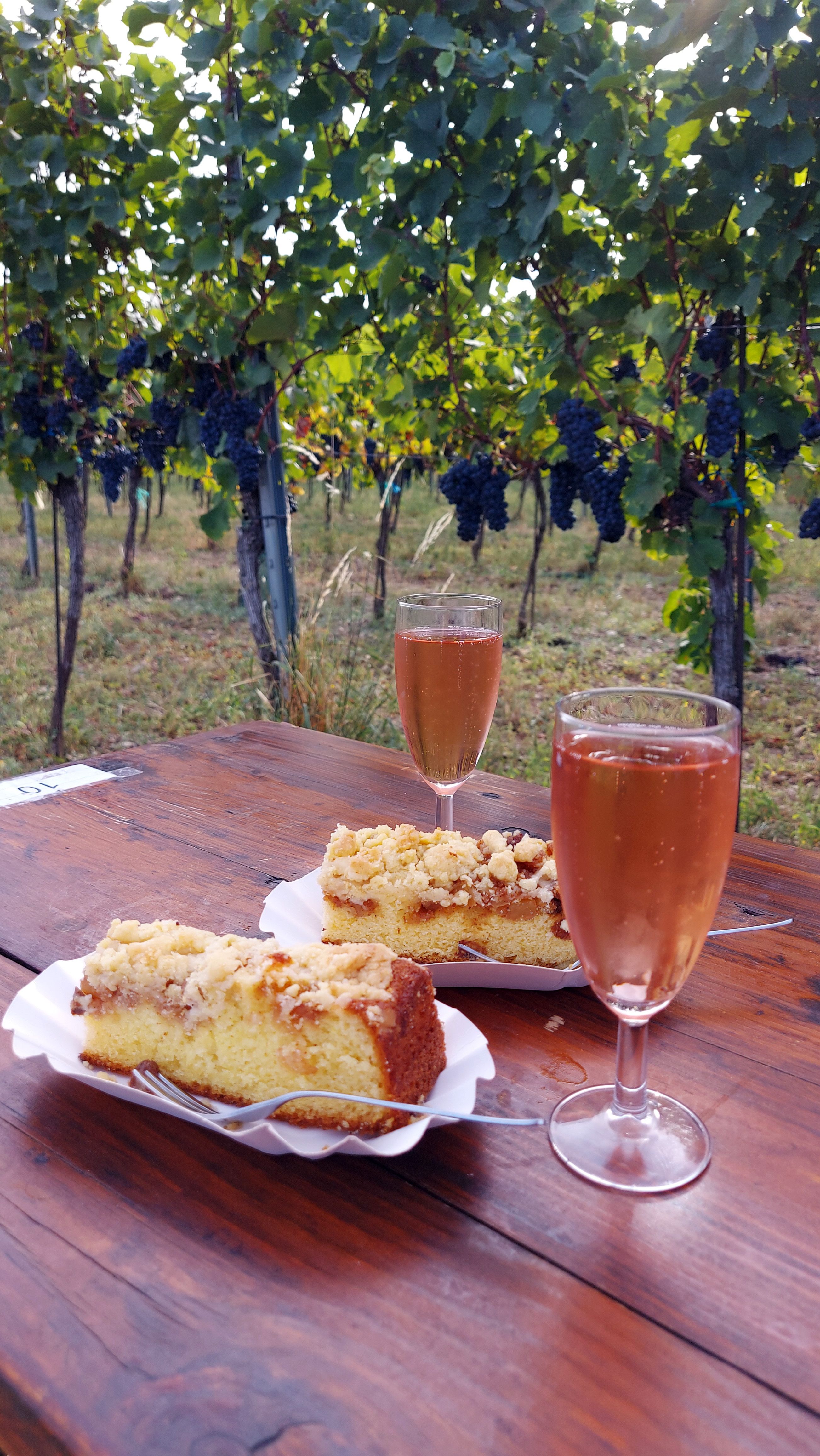 2 Frizzante Gläser und Streuselkuchen stehen auf einem braunen Holztisch im Weingarten bei der Genussmeile