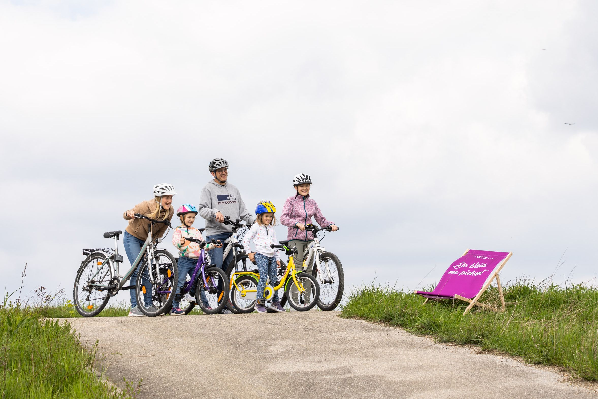 Eine fünfköpfige Familie, bestehend aus zwei Erwachsenen und drei Kindern, fährt mit Fahrrädern fröhlich auf einem Radweg, der sich entlang einer saftig grünen Wiese erstreckt.