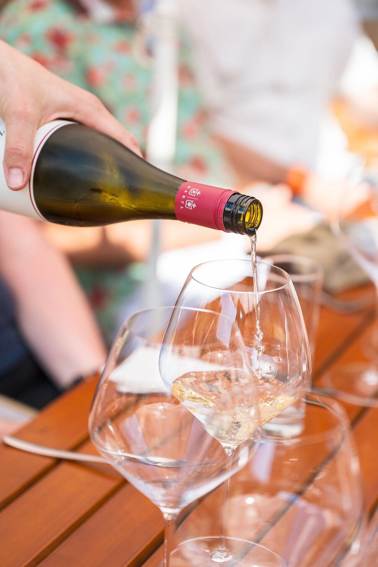 Mit einer Hand wird an einem sonnigen Sommertag weißer Wein in ein Weinglas eingeschenkt; daneben steht ein zweites, leeres Glas bereit.