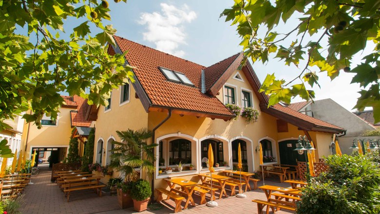 Ein gelbes Gebäude mit roten Dachziegeln und einem Biergarten mit Holzbänken und Sonnenschirmen.