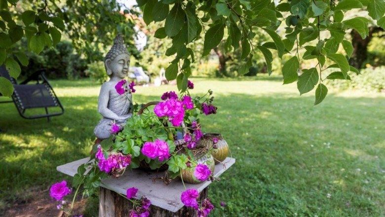 Garten mit Buddha-Statue und pinken Blumen auf einem Holzstumpf.