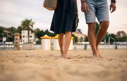Ein Paar streift barfuß durch den Sand im Thermalstrandbad Baden, die Schuhe schwingen lässig in der Hand. Ein Augenblick voller Leichtigkeit, der die Magie eines sommerlichen Abends im Wienerwald einfängt.