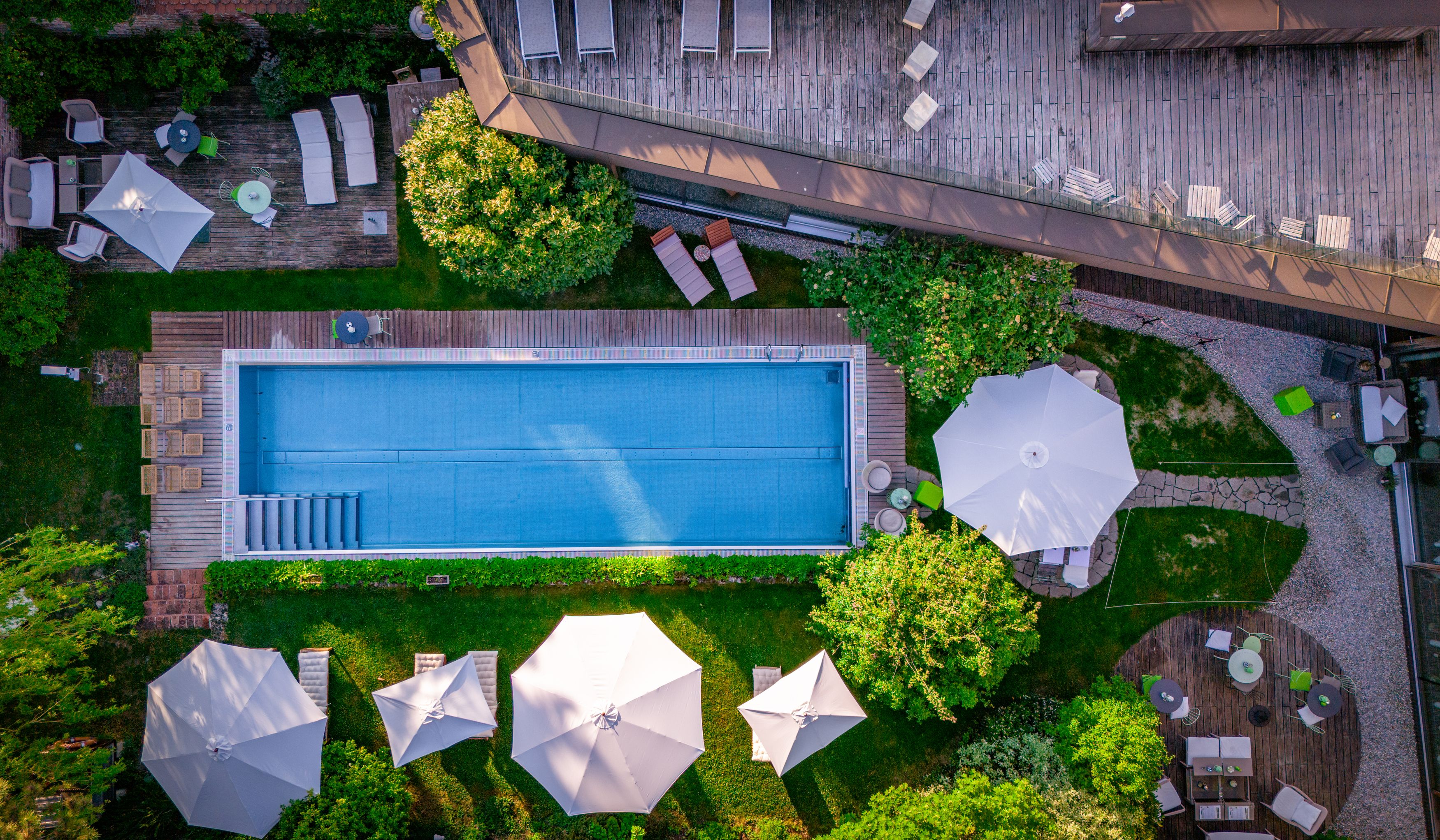 Luftaufnahme eines Pools mit umliegenden Liegestühlen und Sonnenschirmen in einem Garten.
